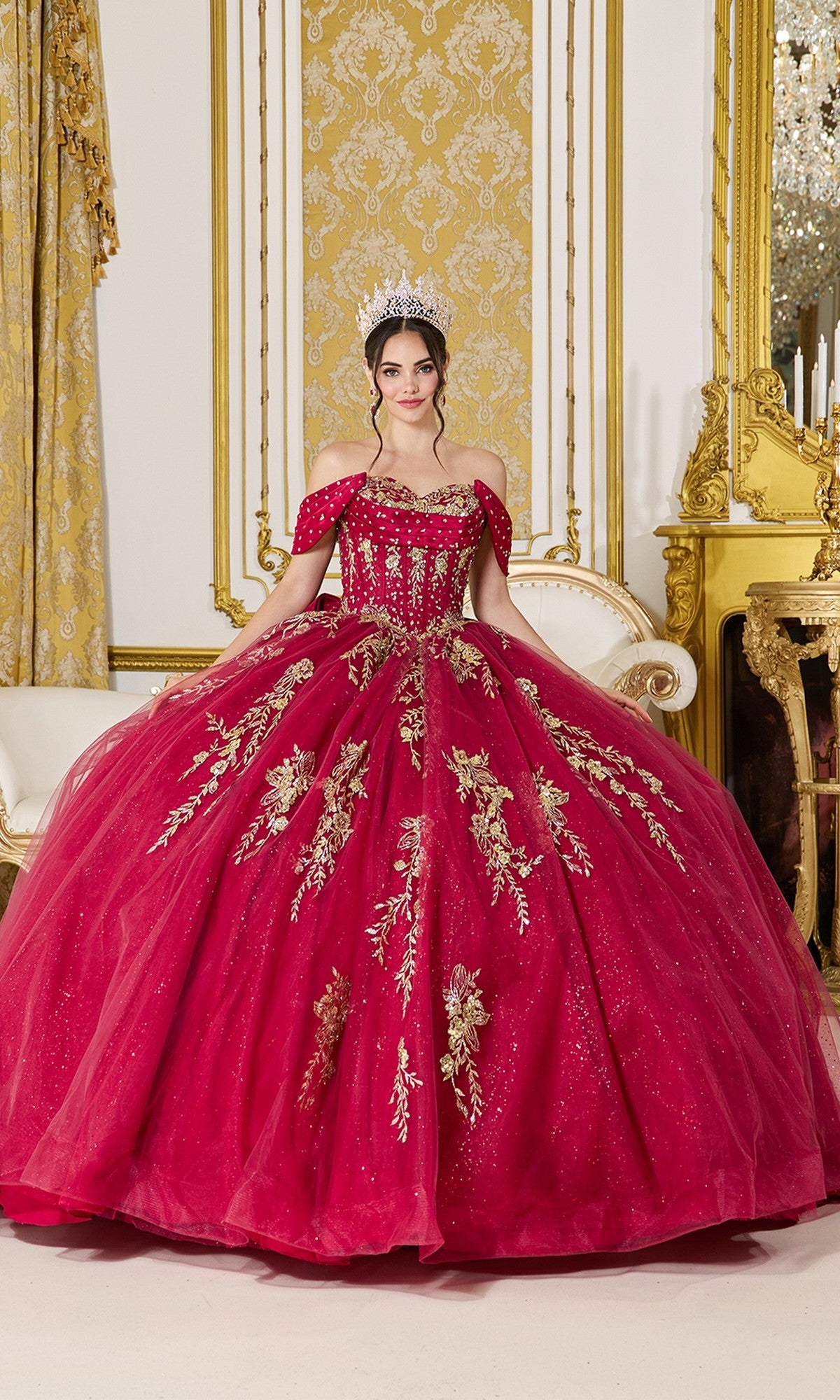 A woman poses indoors in the Quinceanera Dress 5067 by Dancing Queen, a burgundy off-the-shoulder ball gown with gold floral embroidery, against an opulent gold and white background.